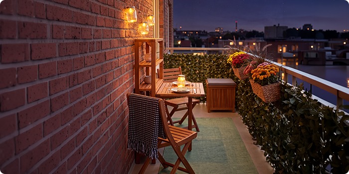 Liten balkong med trestoler og bord, opplyst av lykter og stearinlys, pyntet med blomster i potter og grønne planter, med utsikt over byens silhuett om natten.